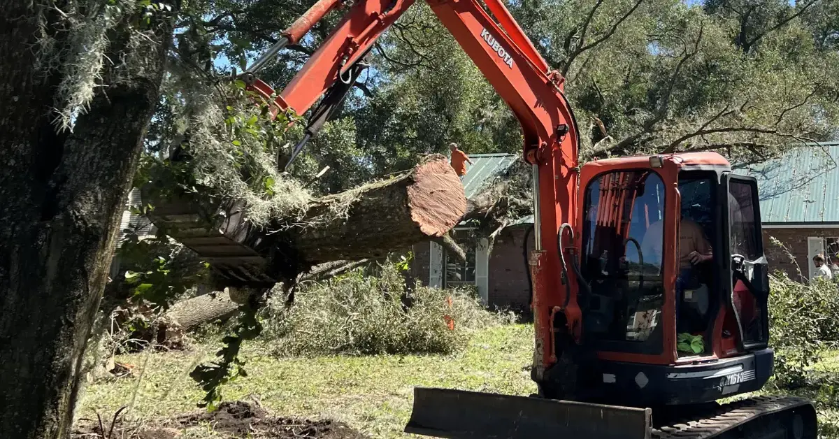 A construction excavator lifting a large cut tree trunk in a yard, with branches and debris scattered near a house in the background.